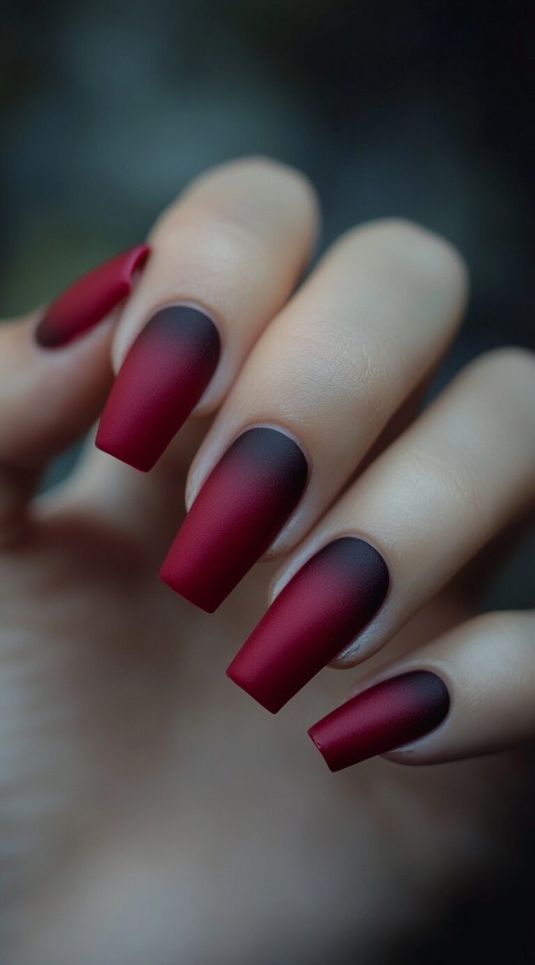 A hand with long coffin-shaped acrylic nails featuring a matte burgundy to black ombre gradient.