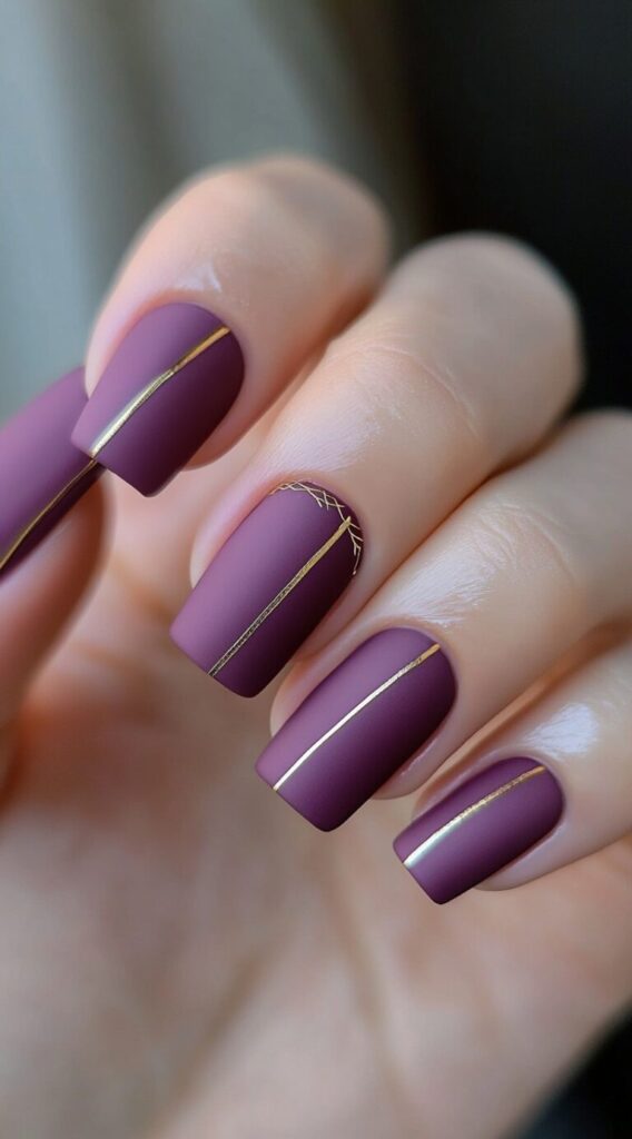 Matte plum square nails decorated with thin gold vertical striping lines, one nail accented with crosshatch gold detail.