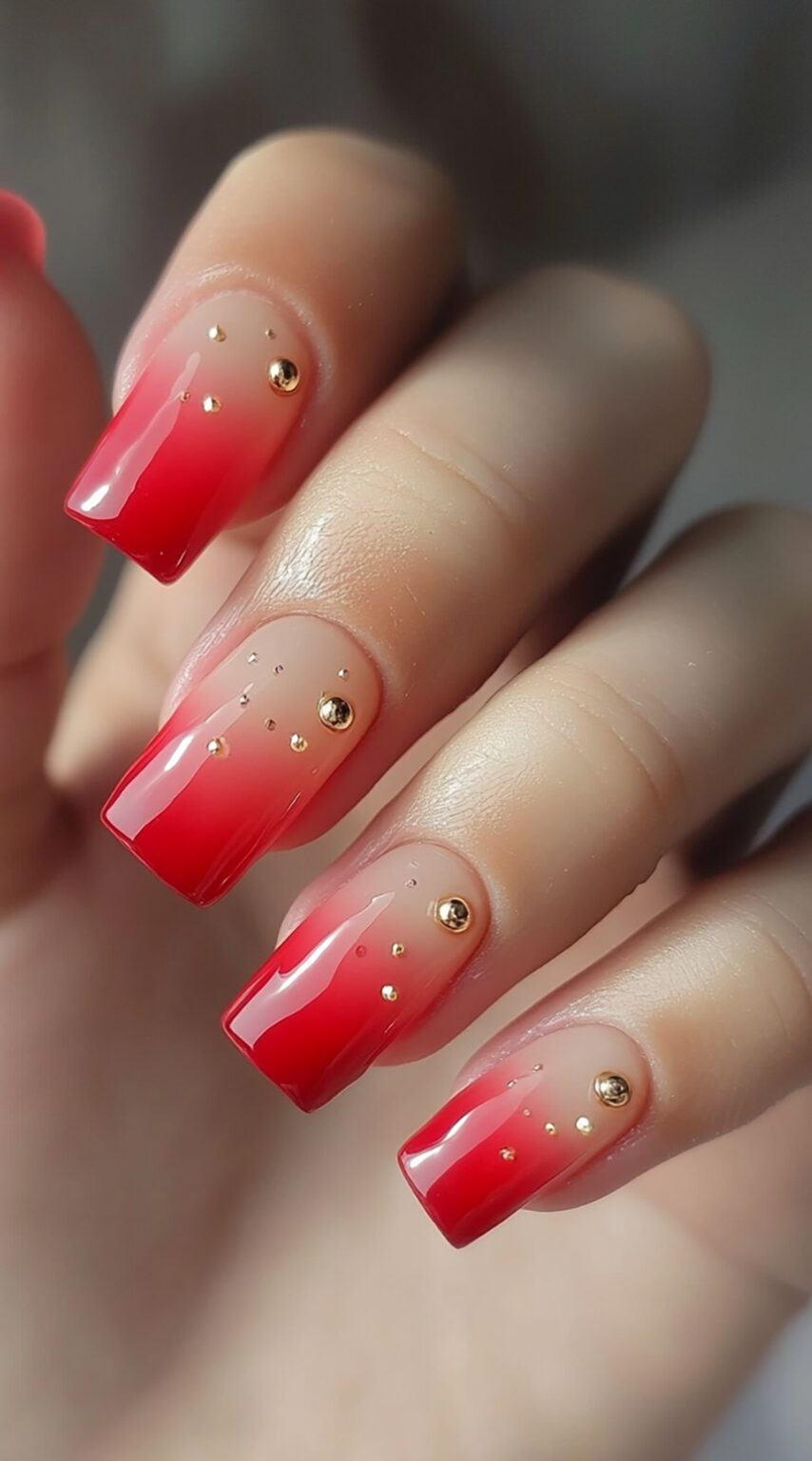A set of red ombre square-shaped acrylic nails with small reflective gold studs near the cuticles.