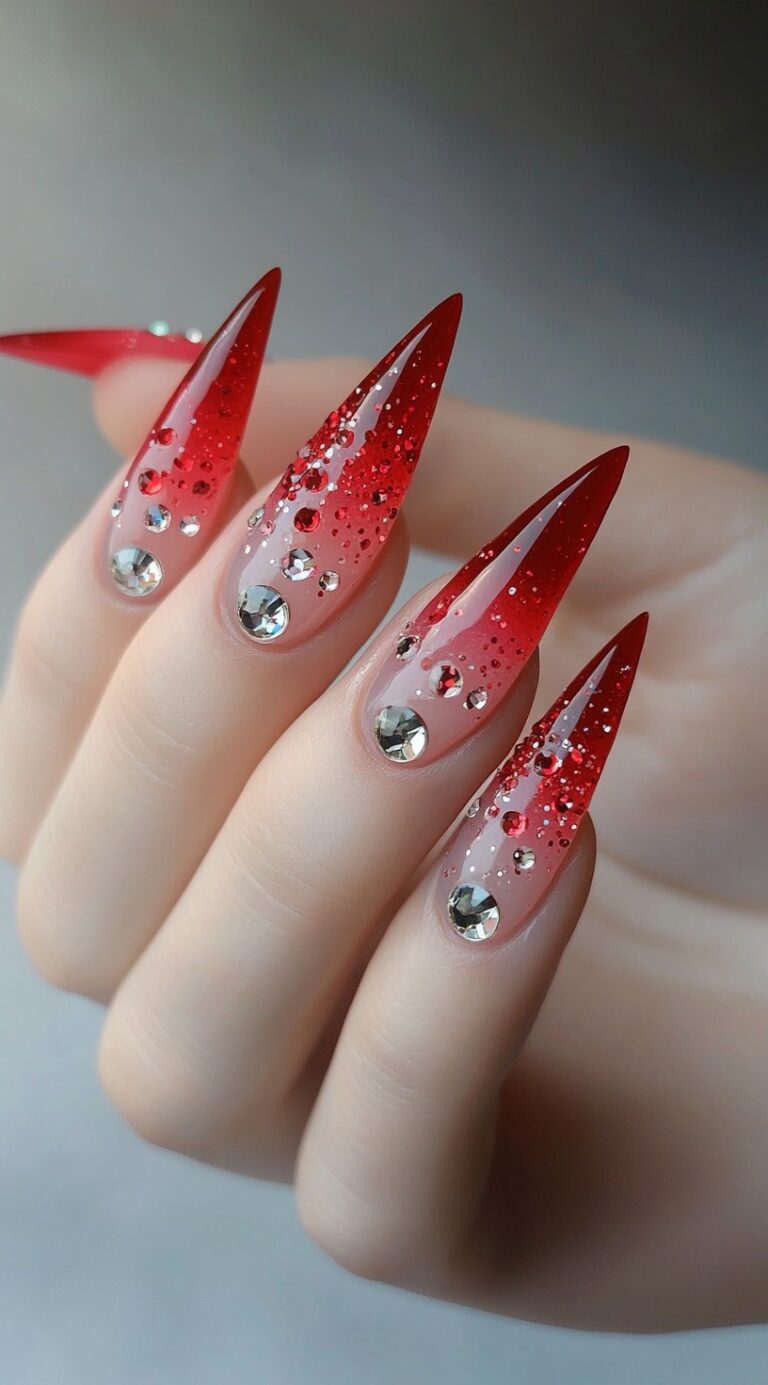 Sharp stiletto nails with a red ombre fade, red glitter, and large rhinestone accents on each nail.